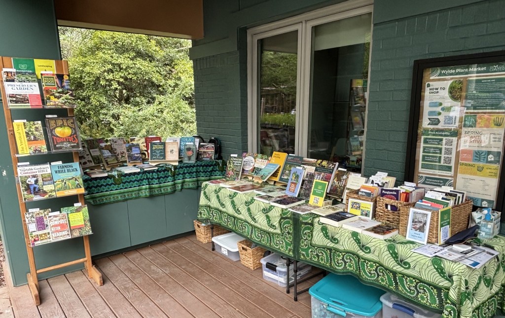 A display of books on gardening and plant care, featuring a wooden bookshelf and a table covered with a green patterned cloth, located outside a building surrounded by greenery.